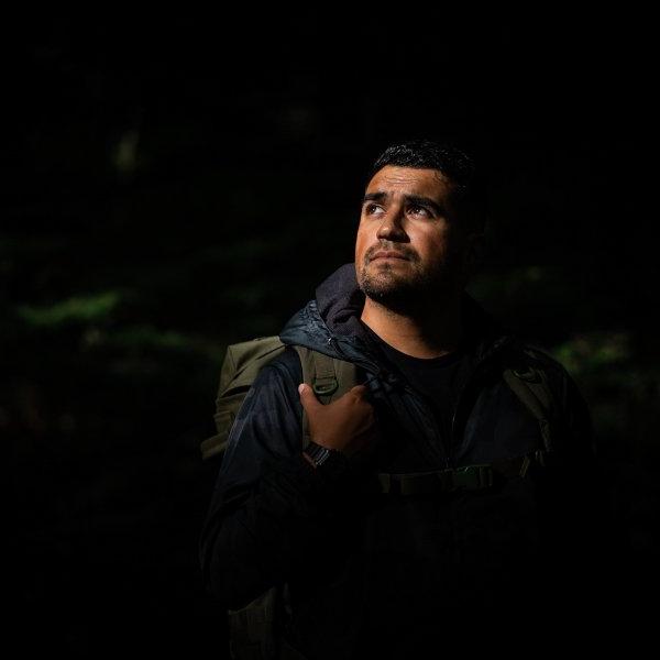 A person stands in the woods holding a backpack as the light hits their face.